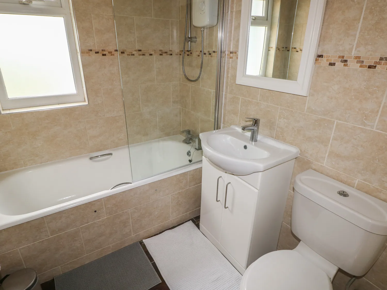 A bathroom with a toilet, a sink with a cupboard below, and a bath with a glass screen and over-bath shower. There is also a mirror above the sink and small window.