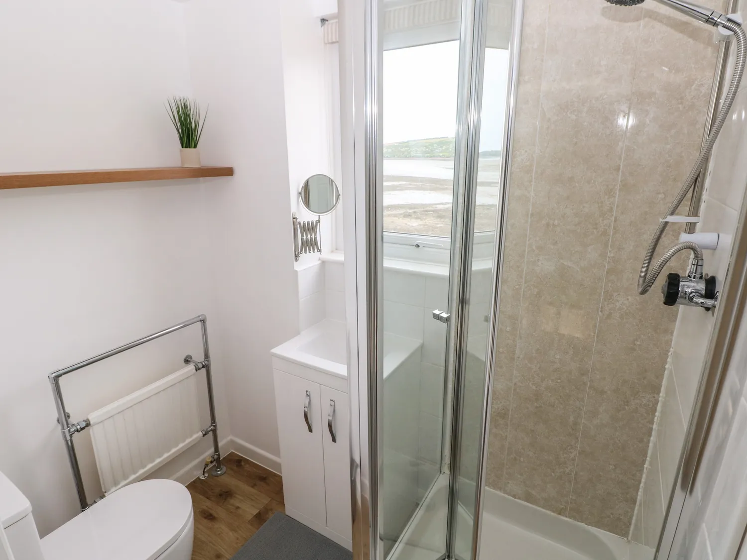 A bathoom containing a walk in shower with a glass door and screen, a sink with a cupboard below, and a toilet. Above the sink is a small mirror and a window. There is also a small radiator.