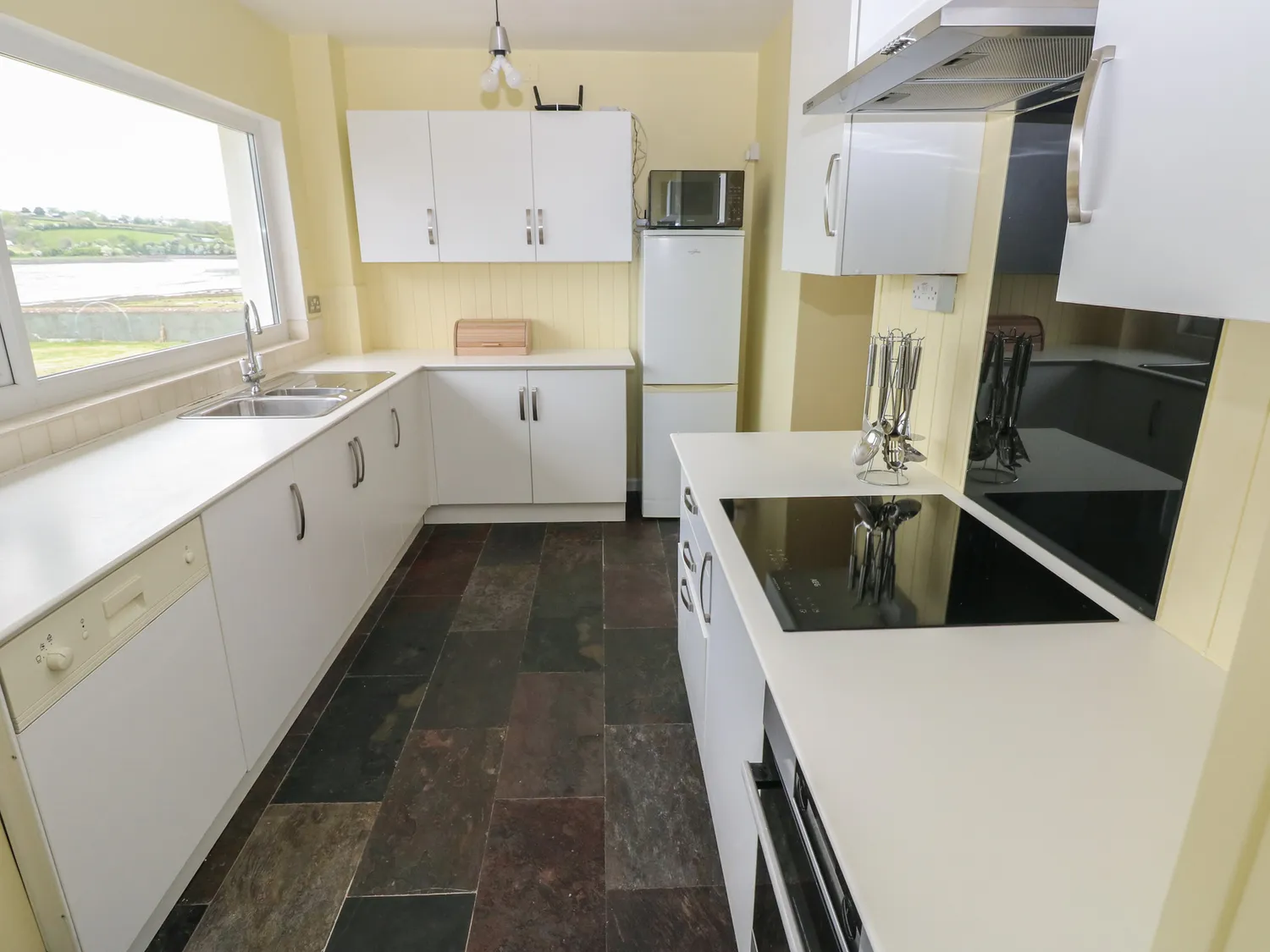 A kitchen with an oven, hob and extractor fan on the right, a dishwasher and sink on the left, and a fridge/freezer with a microwave on top at the back of the room. There are also many under and above counter cupboards.