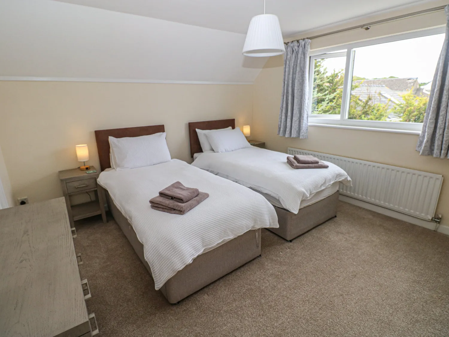 An upstairs bedroom with two single beds next to eachother. Each has its own bedside drawers with a lamp on top. There is also a large window and radiator.