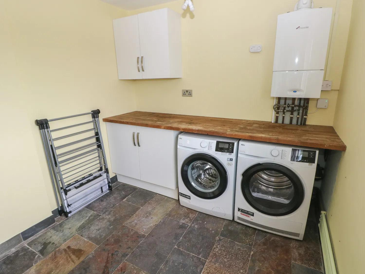 A room with a wooden counter going across a wall. Under the counter are two cupbaords, a washing machine and a dryer. There are also two above counter cupboards and a clothes horse.